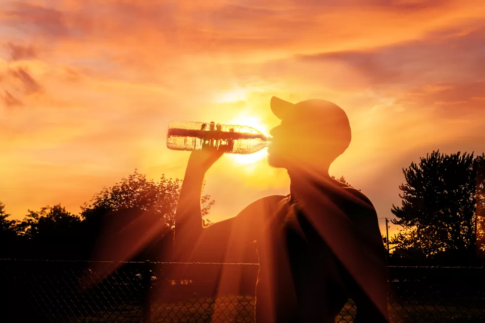 Man drinking water from plastic bottle sillhouetted against orange sky signaling extreme heat